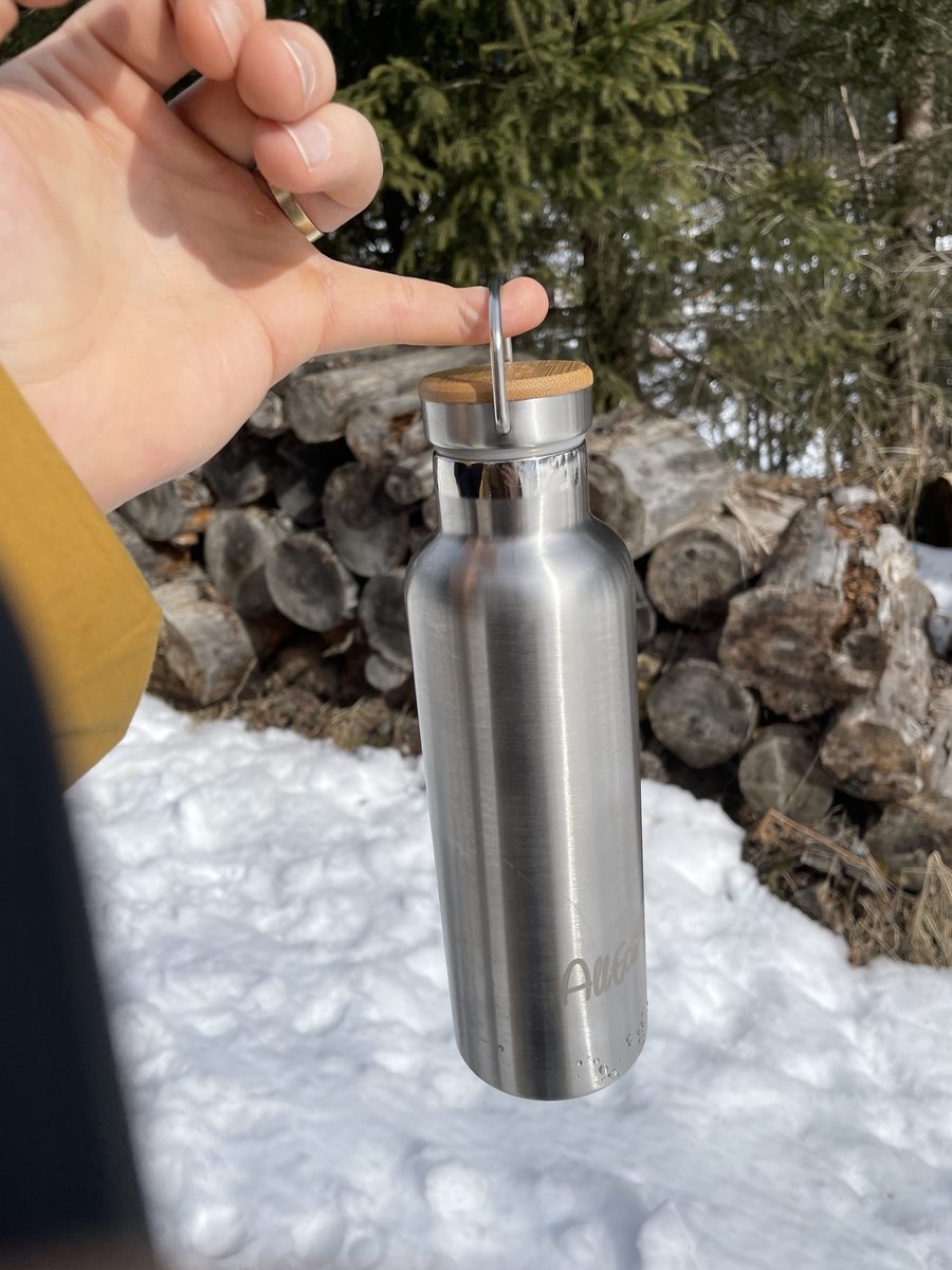 Hand hält eine Edelstahl-Trinkflasche am Tragebügel des Bambusdeckels, im Hintergrund Schnee und Brennholz