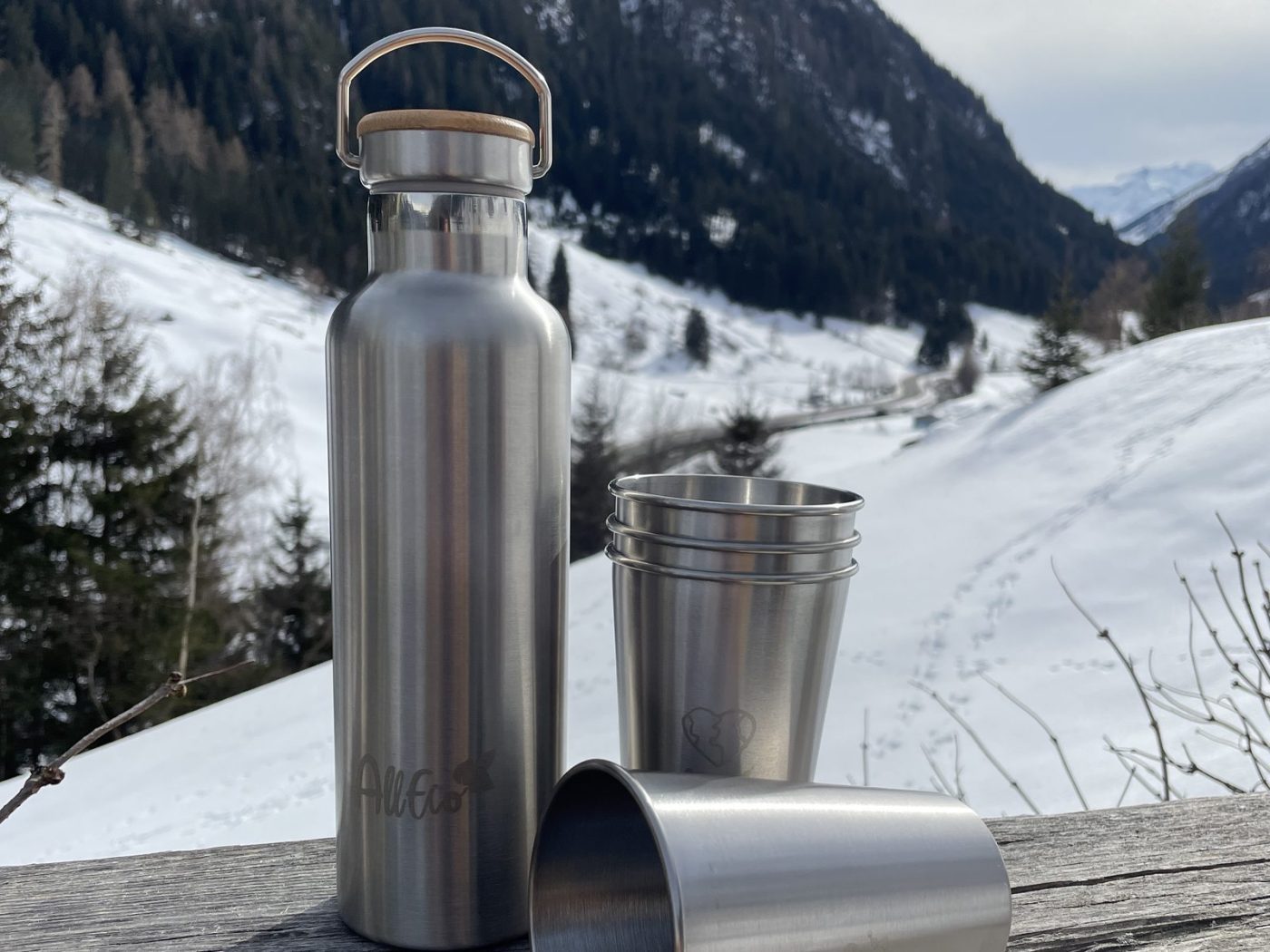 Edelstahl-Trinkflasche von AllEco mit Edelstahlbechern auf einem Holzgeländer vor verschneiten Bergen