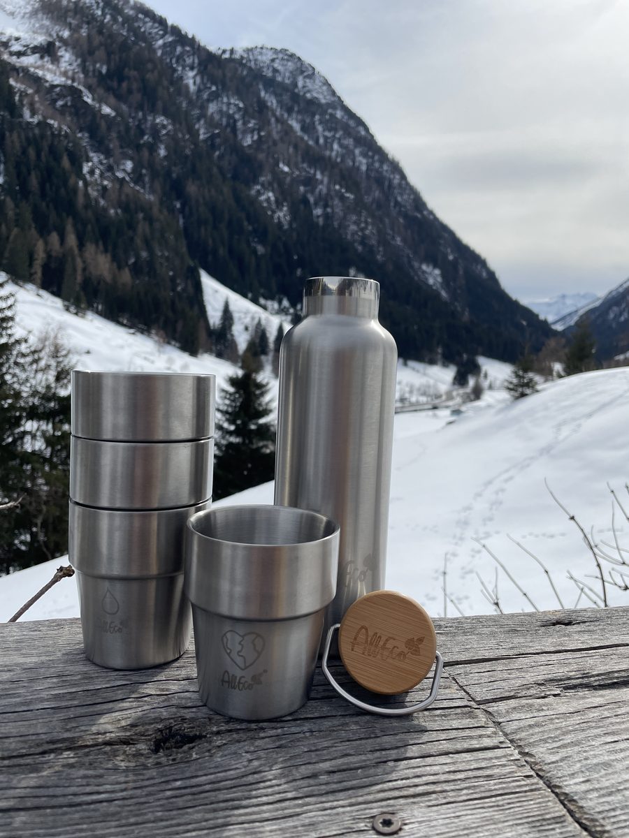 Gestapelte Edelstahlbecher, Trinkflasche und Bambusdeckel auf einem Holzgeländer vor Bergen